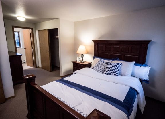Cozy bedroom here at Birchwood Apartments with a dark wood bed frame, white and blue bedding, and patterned pillows. A lit lamp on a bedside table adds warmth. Closet door ajar.