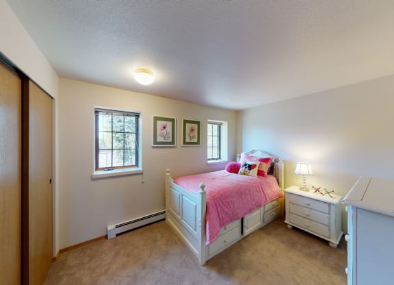 Cozy bedroom here at Birchwood Homes Apartments with a white daybed covered in a pink comforter and colorful pillows. Soft lighting from a lamp. Two floral pictures on the wall.