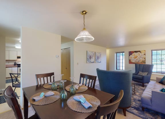 Dining room view of a cozy home with a dark wood table set for four, elegant place settings, and a bright living area featuring plush blue chairs and large windows. Birchwood Homes Apartments.