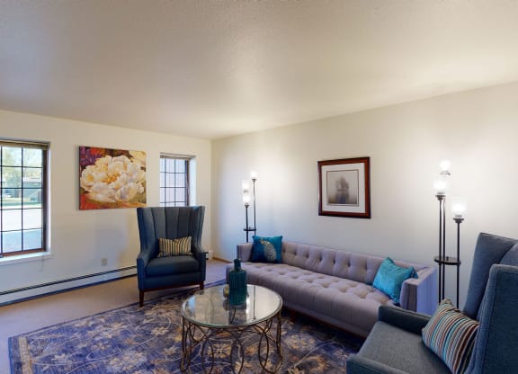 Bright living room here at Birchwood Apartments with modern design; features a gray tufted sofa, blue armchairs, floral artwork, and floor lamps. Large windows illuminate the space.
