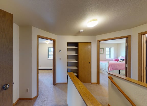 Upstairs hallway here at Birchwood Homes Apartments with beige carpet and white walls, leading to three rooms. Wooden railings and doors add warmth. Bright, simple, and cozy ambiance.