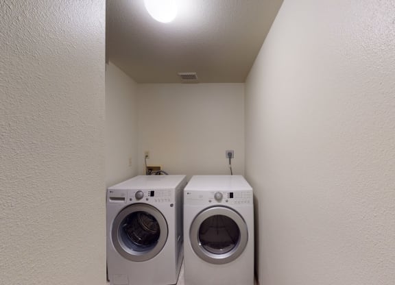 Compact laundry room here at Birchwood Homes Apartments with white walls, featuring a front-loading washer and dryer side by side under a bright ceiling light, conveying a clean, functional space.