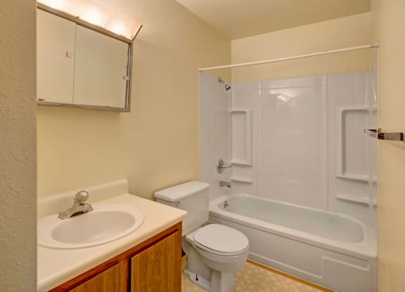 Compact beige bathroom here at Brighton Woods Apartments with a white bathtub, showerhead, toilet, and sink with a mirror above. Simple, clean, and functional design.
