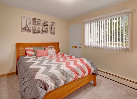 Cozy bedroom here at Brighton Woods Apartments with a wooden bed, chevron-patterned bedding in gray, white, and pink. Sunlight filters through vertical blinds on a window, brightening the space. Black and white art above the bed adds a modern touch.