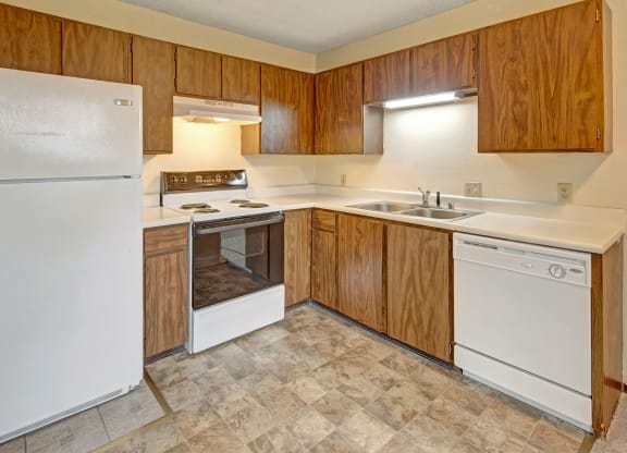Simple kitchen here at Brighton Woods Apartments with wooden cabinets, white appliances including a fridge, stove, and dishwasher. Neutral tones create a clean, functional space.