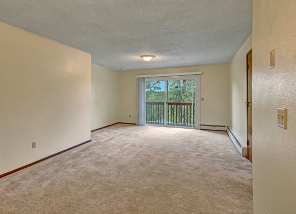 Spacious empty living room here at Brighton Woods Apartments with beige carpet, cream walls, a central ceiling light, and sliding glass doors leading to a balcony with a green view.