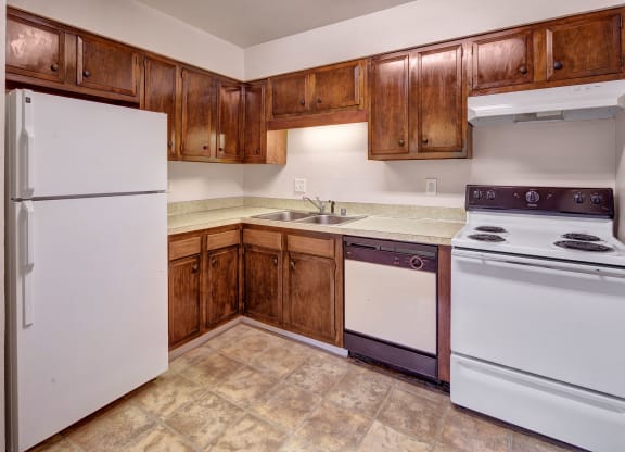 Compact kitchen here at Castle Apartments with wooden cabinets, white fridge, oven, and dishwasher. Neutral tones, tiled floor, and bright lighting create a clean, simple feel.