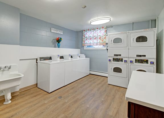 Laundry room here at Century Plaza with a cozy feeling. Features four white washers, stacked dryers, a utility sink, floral curtain, wood floor, and decorative flowers.