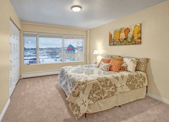 A cozy bedroom here at Century Plaza with beige walls and carpet, featuring a bed with patterned bedding and colorful cushions. Large window shows a snowy view outside.