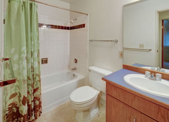 A clean, tidy bathroom here at Chugach South Apartments with a light green shower curtain featuring leaf patterns, a white bathtub, a toilet, and a wooden vanity with a blue countertop.