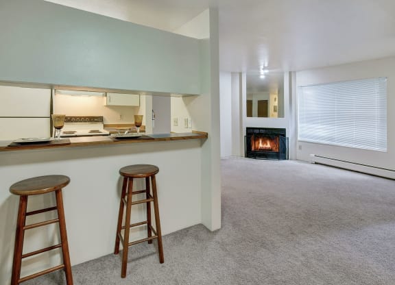 Spacious, cozy living area here at Chugach South Apartments with a grey carpet, featuring a kitchen bar with two wooden stools on the left, and a lit fireplace on the right.
