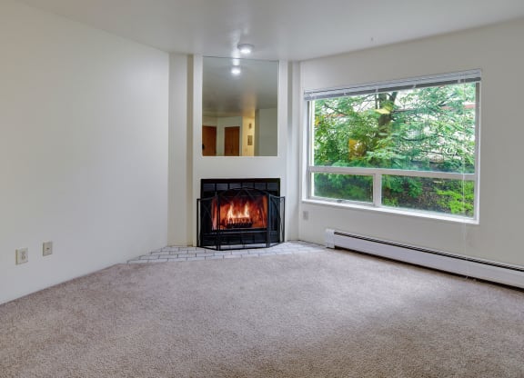 A cozy room here at Chugach South Apartments with a lit fireplace, beige carpet, and a large window showing green foliage. The atmosphere is warm and inviting, ideal for relaxation.