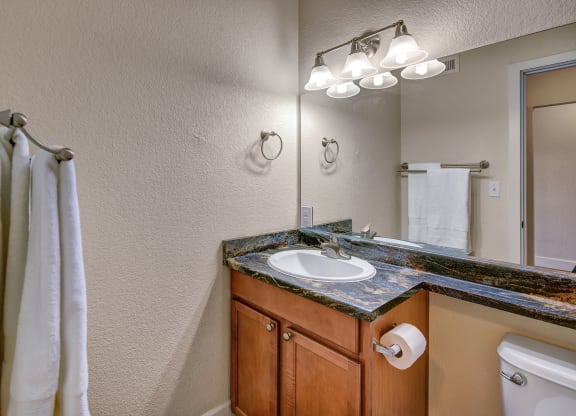 Bathroom here at City View Apartments with wooden vanity, marble countertop, and mirror. Three-light fixture above, white towels on racks, and a roll of toilet paper. Clean and modern.