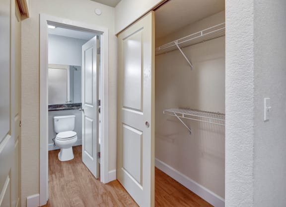 Hallway here at City View Apartments showing an open closet with wire shelves to the right, and a view into a bathroom with a white toilet and mirror on the left. Wood flooring.