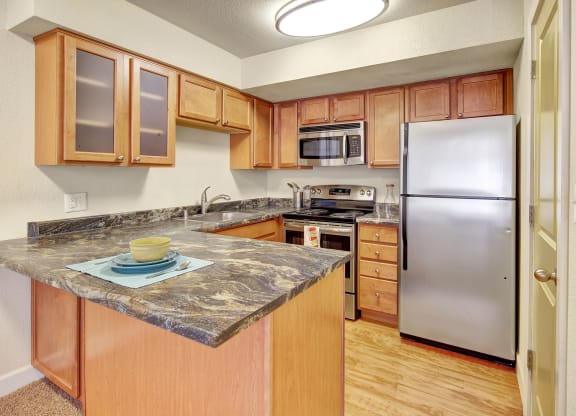 Compact kitchen here at City View Apartments with light wood cabinets and marble countertops. Stainless steel appliances, including a fridge and microwave. Cozy and inviting feel.