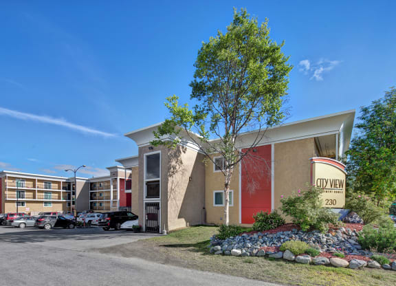City View Apartment complex under a clear blue sky, featuring modern beige buildings with red accents, a parking lot with cars, and landscaped greenery.