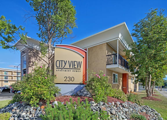 Apartment complex exterior with a sign reading "City View Apartment Homes 230." Surrounded by trees and shrubs, under a clear blue sky. Inviting and serene.
