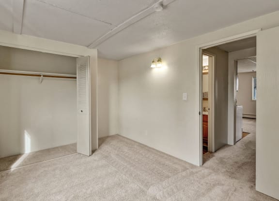 Empty beige carpeted bedroom here at College View Apartments with an open closet on the left, showing hanging rod, and an open door leading to a hallway. Dim wall light creates a cozy feel.