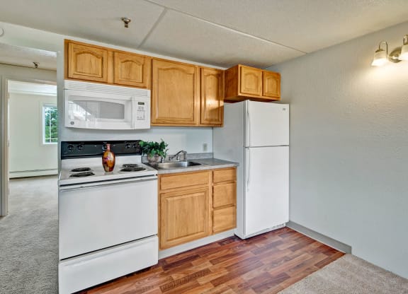 Compact kitchen here at College View Apartments with wooden cabinets, white appliances, and a vase on the stove. Bright room with hardwood floors, adjacent to a carpeted space.