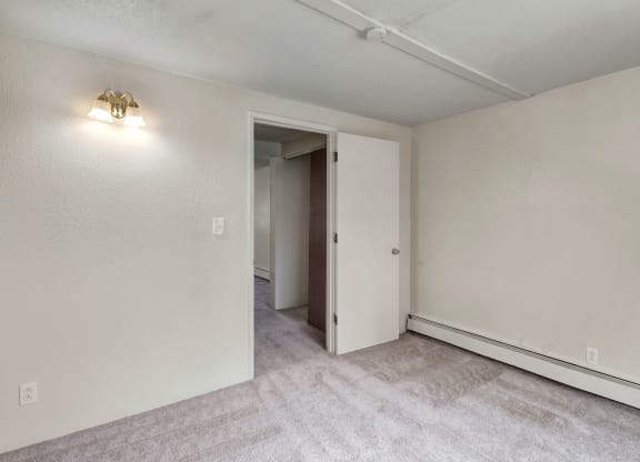Empty bedroom here at College View Apartments with beige walls, light gray carpet, a closed white door slightly ajar, and a wall sconce casting soft light, creating a neutral tone.