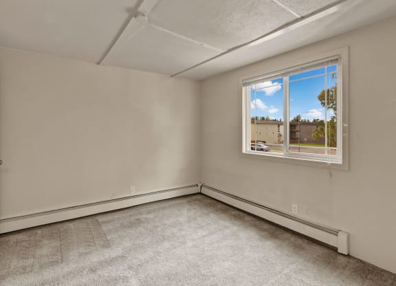 Empty bedroom here at College View Apartments with beige walls, light gray carpet, and a large window revealing a view of trees and buildings outside. The tone is calm and neutral.