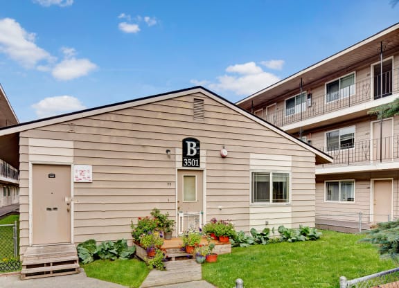 College View Apartments exterior of a beige building labeled "B 3501" with potted plants and flowers in front. Surrounding buildings have balconies. Clear blue sky above.