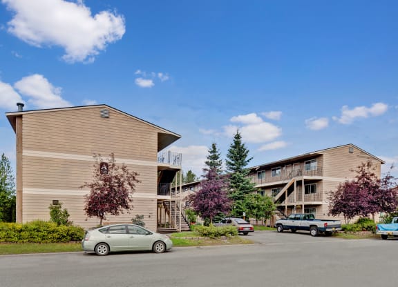 Two beige apartment buildings here at College View Apartments with wooden siding, surrounded by trees and parked cars under a clear blue sky. The scene conveys a peaceful suburban setting.