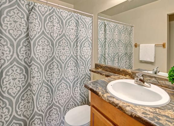 Bathroom here at Conifer Grove Apartments with a decorative green and white patterned shower curtain, a brown marble countertop, green potted plants, and a white towel on a rack.