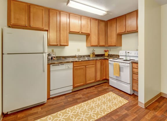 A cozy kitchen here at Conifer Grove Apartments with warm wooden cabinets, white appliances, dark countertops, and a patterned yellow rug on hardwood flooring, creating a welcoming atmosphere.