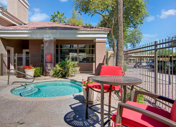 Small hot tub in a sunlit courtyard here at Dobson Towne Centre Apartments, surrounded by a brown fence and a building. Red chairs and table add a vibrant touch. Relaxing atmosphere.