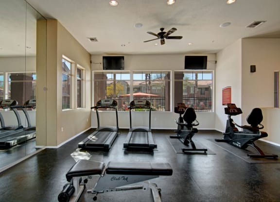 A bright gym here at Dobson Towne Centre Apartments with two treadmills and two recumbent bikes facing large windows. A weight bench is in the foreground; the atmosphere is clean and inviting.