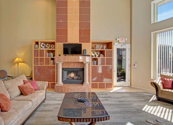Spacious resident lounge here at Dobson Towne Centre Apartments with beige walls, tall fireplace, and wooden shelves. Cozy beige sofa with red cushions. Sunlight streams through large windows. Relaxed, inviting ambiance.