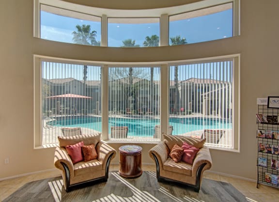 Cozy seating area here at Dobson Towne Centre Apartments with two beige armchairs and a round table, facing large windows overlooking a sunny pool, creating a relaxed and inviting atmosphere.