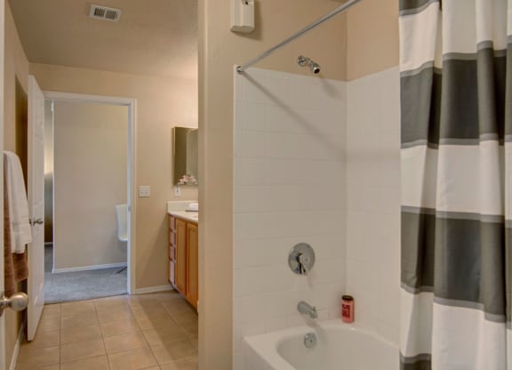 Bathroom here at Dobson Towne Centre Apartments with a white bathtub, gray-striped shower curtain, and white tiles. Beige walls lead to a hallway with a white chair visible. Cozy ambiance.
