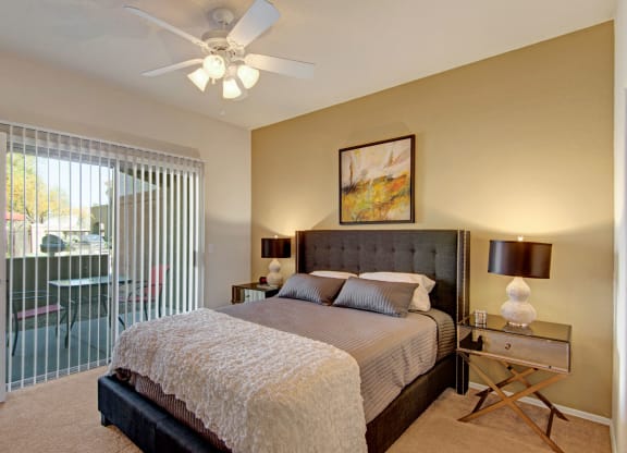 Modern bedroom here at Dobson Towne Centre Apartments with a gray upholstered bed, striped bedding, and a textured white throw. Warm lighting from side lamps, abstract art above bed, and sliding glass door with vertical blinds leading to a patio.