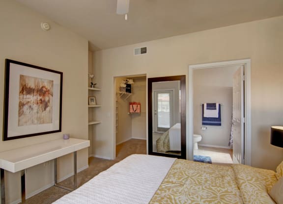 Modern bedroom here at Dobson Towne Centre Apartments with beige walls, white desk, abstract art, and a large mirror. Walk-in closet and bathroom visible, creating a cozy, organized feel.