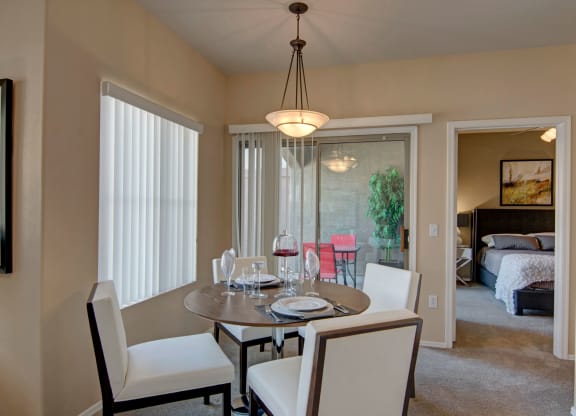 Dining area here at Dobson Towne Centre Apartments with a round table set for four. White chairs and a modern chandelier create a cozy atmosphere. A glass door leads to a patio, and a bedroom is visible.
