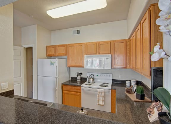 Cozy kitchen here at Dobson Towne Centre Apartments with light wood cabinets, white appliances, and a granite countertop. A kettle sits on the stove; orchids and decor add warmth.