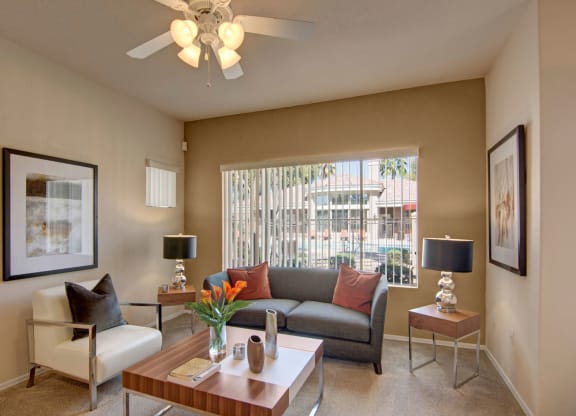 Modern living room here at Dobson Towne Centre Apartments with a gray sofa and white armchair featuring orange pillows. A small coffee table holds a vase with flowers. Large window with blinds lets in sunlight. Two framed pictures and stylish lamps add elegance. Airy and inviting.