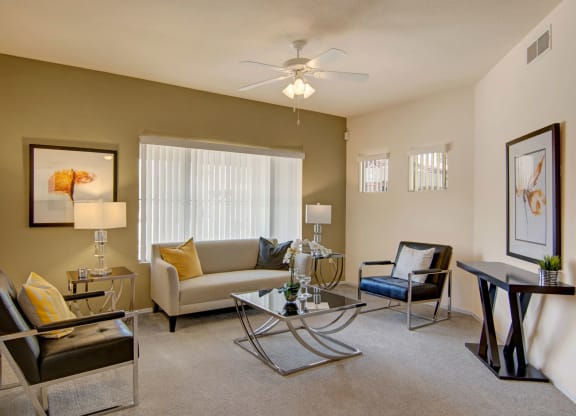 Modern living room here at Dobson Towne Centre Apartments with a neutral tone, featuring a beige sofa, two black chairs, glass coffee table, abstract art, and natural light from large windows.