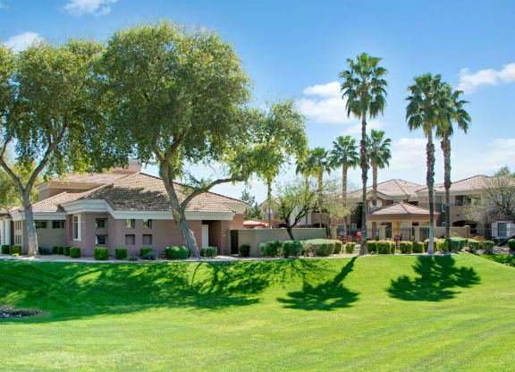 A serene suburban scene here at Dobson Towne Centre Apartments with a tan stucco house surrounded by lush green lawns, palm trees, and clear blue skies. Shadows create a tranquil atmosphere.