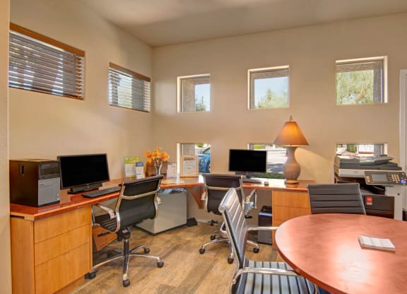 Modern office space here at Dobson Towne Centre Apartments with neutral tones, featuring two desks with computers, black chairs, a lamp, and a printer. Natural light streams through small windows.