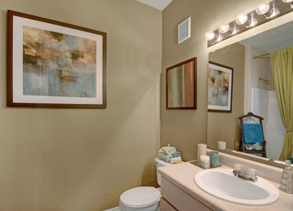 A cozy bathroom here at Pala Mesa Apartments with beige walls features abstract art, a wide mirror with bright lights, and neatly arranged towels. A shower curtain adds a touch of green.