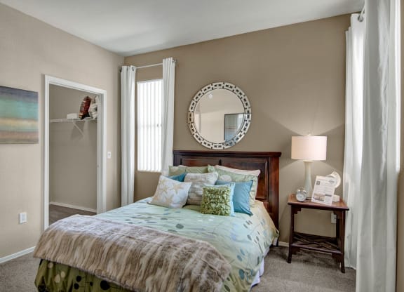 A cozy bedroom here at Pala Mesa Apartments with a wooden bed adorned with blue and green pillows. A circular mirror hangs above the headboard. Soft light from a table lamp on a wooden nightstand creates a warm ambiance. Light tan walls and white curtains enhance the serene atmosphere.