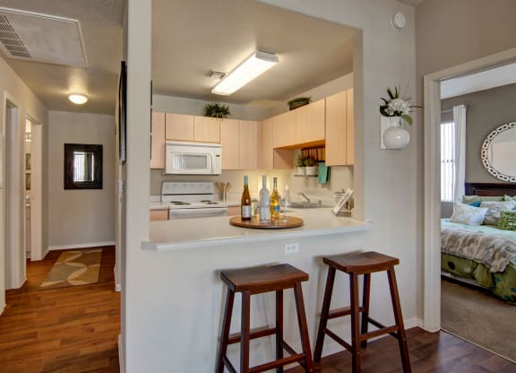 Compact kitchen here at Pala Mesa Apartments with light wood cabinets, a white stove, and microwave. Counter with wine bottles and glasses, two wooden stools. Cozy, inviting feel.