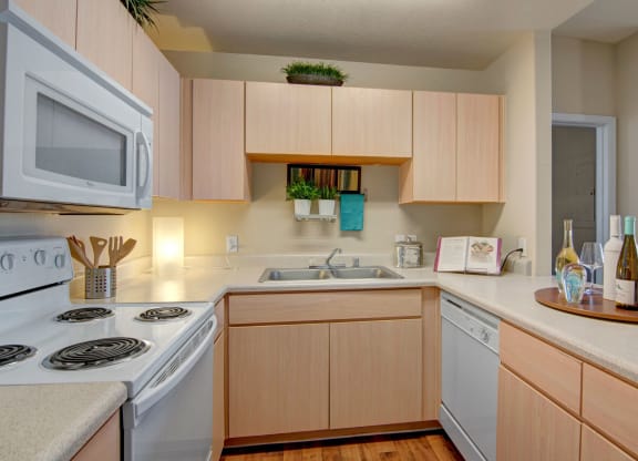 Modern kitchen here at Pala Mesa Apartments with light wood cabinets, beige countertops, and appliances including a microwave and stove. Decorative plants and wine bottles add warmth.