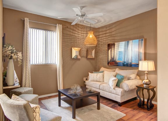 Cozy living room here at Pala Mesa Apartments with cream sofa, teal and patterned pillows, brown coffee table, floral vase, and lamp. Warm lighting and a serene ocean painting.