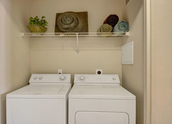 Compact laundry room here at Pala Mesa Apartments with a washer and dryer side by side. Above, a shelf holds a potted plant, three rolled towels, and spiral decor. Neutral tones.