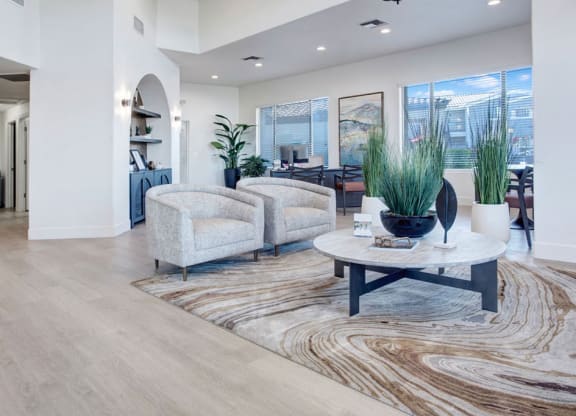 A modern, spacious resident lounge here at Pala Mesa Apartments with light wood flooring and two beige armchairs around a round marble table. Large plants, abstract art, and soft lighting create a tranquil, inviting atmosphere.