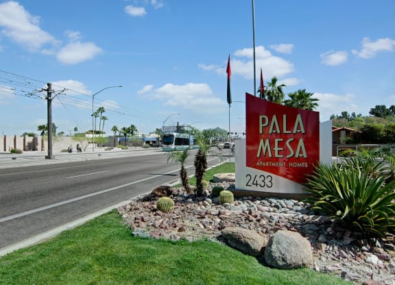 Landscape view of Pala Mesa Apartment Homes sign with palm trees and desert plants. A streetcar passes by on a sunny day, blue sky with clouds.